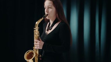 Siyah elbiseli genç bir kadın stüdyoda siyah arka planda saksafon çalıyor. Saksafon çalan caz müzisyeni. Siyah bir gece elbisesi giymiş, sahnede solo yapan bir kadın saksafoncu..