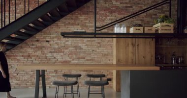 Kadın, duvarları tuğlalı ve siyah merdivenli modern ve geniş ahşap mutfak odasında sandalyede oturuyor. Gayrimenkul. Ahşap adası ve sandalyeleri olan lüks bir mutfak. Gerçek Lüks Tasarım İçi.