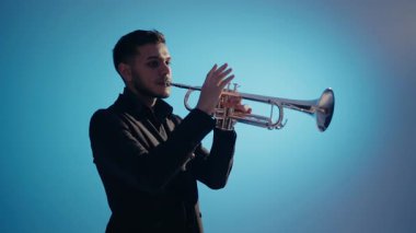 Takım elbiseli genç ve yakışıklı bir adam arzu ve ilhamla trompet çalar. Modern bir stüdyoda mavi arka planda trompet çalan bir erkek caz şarkıcısı. Canlı caz konseri..