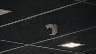 Güvenlik kamerası bir tavana monte edilmiş, güvenlik ve gözetim sağlanıyor. Kamera lensi tavan kiremitleri ve aydınlatma armatürlerinin arka planında belirgin bir şekilde gösteriliyor. Bölgeyi gözlüyor.