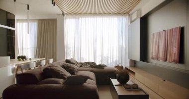 Modern oturma odası içi rahat kahverengi kanepe, şık ahşap tavan ve katıksız perdeli geniş pencere. Doğal ışık samimi ortamı güçlendirir..