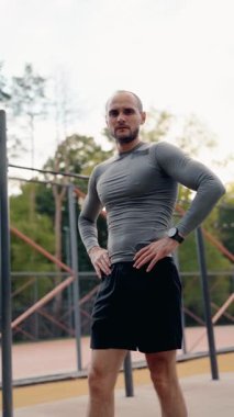 Formda bir adam dışarıda çalışmaya ara verir. Elleri kalçasında duruyor, soluklanıyor. Gri bir tişört ve siyah şort giyiyor..