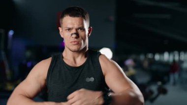 Kaslı bir adam spor salonunda pazılarını esnetir. Siyah bir atlet giyiyor ve burnunda bir bandaj var. Kararlı ve güçlü görünüyor. Bu video fitness ve vücut geliştirme için mükemmel.,