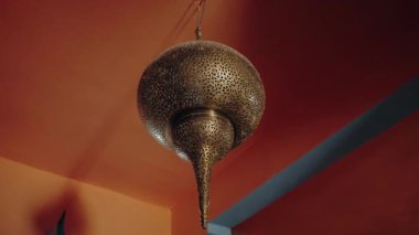 Süslü Fas pirinci tavan lambası tavandan sarkıyor. Karmaşık, delinmiş metal kalıplar turuncu-kırmızı duvarlar ve gök mavisi aksanlar üzerinde ışık desenleri oluşturur. Zarif ve geleneksel tasarım