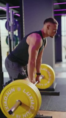Kaslı bir adam, mor ışıklandırmalı modern bir spor salonunda sarı ağırlıklarla kaldırma yapıyor. Uygun forma ve tekniğe odaklanıyor. Bu video güç, uygunluk ve kararlılığı somutlaştırır.,