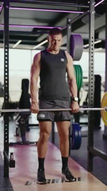 Kaslı bir adam modern bir spor salonunda asansör kaldırır. Ağırlık kaldırıyor, dayanıklılık ve zindelik gösteriyor. Spor salonunun mor neon ışıkları ve çeşitli spor aletleri var. Sağlık, sağlık ve adanmışlık.