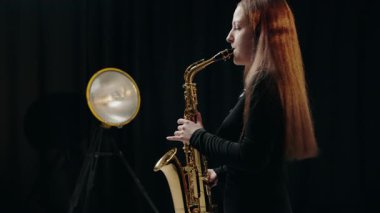 Siyah elbiseli genç bir kadın stüdyoda siyah arka planda saksafon çalıyor. Saksafon çalan bir bayan müzisyen. Siyah bir gece elbisesi giymiş, sahnede solo yapan bir kadın saksafoncu..