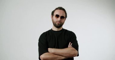 Güneş gözlüklü ve çapraz kollu sakallı bir adam beyaz bir zemine karşı duruyor. Kendine güveni ve tarzı yansıtıyor. Minimalist ortam onun varlığını vurguluyor, yaşam tarzı için ideal hale getiriyor.,