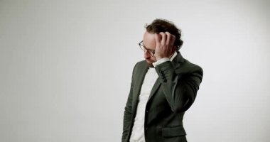 Takım elbiseli ve gözlüklü bir işadamı saçını düzeltir ve beyaz bir arkaplanda gülümser. Bu video güven, profesyonellik, başarı ve stil duyguları uyandırıyor. İş ve şirket için ideal.,