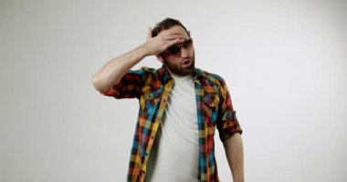 Sakallı ve güneş gözlüklü genç bir adam, beyaz bir arka plana karşı hayal kırıklığı ve inançsızlık ifade ediyor. Elleriyle jest yapıyor ve sinir bozucu ve çileden çıkarıcı yüz ifadeleri yapıyor..