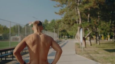 Güneşli bir günde spor sahasının yanında çalışan kaslı bir gencin arka görüntüsü. Sahne, açık hava fitness rutinlerinin enerjisini ve adanmışlığını yakalıyor. Genç Sporcu Adam Egzersiz Yapıyor