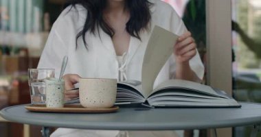 Genç bir kadının, elinde bir fincan kahve ve masanın üzerinde serinletici bir bardak suyla kitaplara daldığı sakin bir sahne. Rahat bir kafe ortamında yakalanmış bir eğlence ve öğrenme anı.