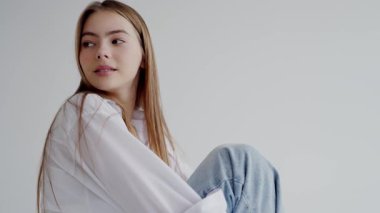 Sarışın, beyaz bluz ve kot pantolon giyen, hafif ışıklandırılmış stüdyo ortamında poz veren genç bir kadının yakın plan portresi. Huzurlu Genç Kadın Bakıyor Yumuşak bir gülümsemeyle