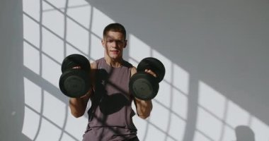 Atletik yapılı bir adam stüdyoda yoğun bir şekilde trenler yapıyor, dambılları gölgeler altında kaldırıyor. Stüdyo Ayarlarında Dambıllarla Atletik Genç Adam Eğitimi