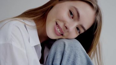 Genç bir kadın bacaklarını kavuşturup yerde oturuyor, gülümsüyor, tek kaşını kaldırıyor ve eliyle şakacı bir jest yapıyor. Genç bir kadın yüzünde bir tebessümle oturuyordu.