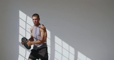 23 yaşında atletik yapılı bir adam iyi ışıklandırılmış bir stüdyoda ağırlık çalışması yapıyor. Güneş Işığı Stüdyosunda Ağırlık Kaldıran Genç Atletik Adam