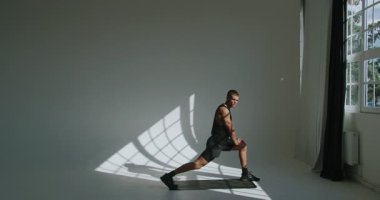23 yaşında atletik bir erkek, stüdyoda esniyor, figürü dramatik bir ışık ve büyük bir pencerenin yanında gölgeler içinde. Dramatik Gölgelerle Parlak Stüdyoda Genç Atletizm Eğitimi