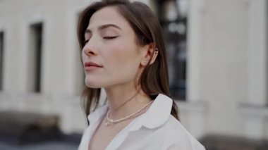 Beyaz bluzlu beyaz bir kadının şehir ortamında kendine güvenen bir şekilde poz verdiği yakın plan bir görüntü. Yüzünde bir gülümseme ifadesi var. Genç Kadın 'ın Yakın Çekim Portresi 