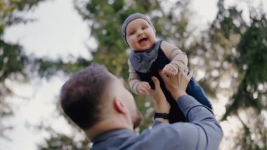 Neşeli bir babanın, yeşil ağaçlarla çevrili ve yaprakların arasından süzülen bebeğini havaya kaldırması ne hoş bir manzara. Neşeli Baba, dışarıda Sevimli Bebek Lisesi 'ni kaldırıyor.