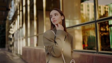 Bej ceketli şık bir genç kadın şehir ortamında güven içinde duruyor. Bakışları yumuşak ve davetkar camdan binalar arasında. Şık Bej Ceketli Zarif Genç Kadın Şehir Merkezi