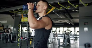 Odaklanmış genç bir adam, sağlık ve dayanıklılığı yansıtan iyi donanımlı bir fitness merkezinde antrenmanlar arasında suyla sulanır. Spor salonunda Antrenman Molasında Su İçen Genç Adam