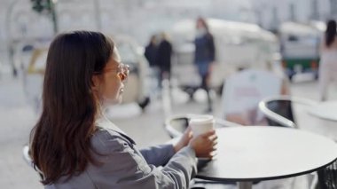 Genç bir kadın bir sokak kafesinde oturuyor, kahvesini yudumluyor ve hareketli şehir hayatını seyrediyor. Şehir ortamında mükemmel bir an. Yoğun Şehir Kafe 'sinde Kahve İçen Genç Kadın