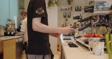 Markalı siyah tişörtlü erkek bir barista çağdaş bir kafe ortamında kaliteli bir espresso makinesi kullanıyor. Görüntü, tam kahve yapma anını yakalıyor. Chisinau - Moldova. 27 Temmuz 2023