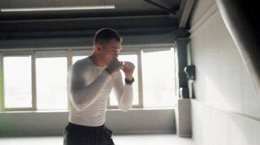 Uzun kollu beyaz gömlekli genç bir atletik parlak bir spor salonunda ağır bir torbaya boks yumrukları uyguluyor. Spor salonunda Kum Torbasıyla Odaklanmış Genç Adam Eğitimi