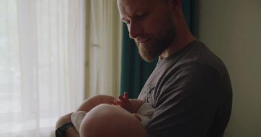 Genç adam baba, bebeği elinde tutuyor, sallıyor ve çocuğu uyutuyor. Hem de çocuk odasında dururken. Mutlu ebeveynlik kavramı