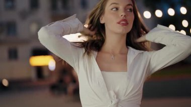 Şık beyaz bir bluz içindeki çarpıcı bir kadın zarif bir şekilde şehir ışıklarının arkasına poz vererek büyüleyici ve büyüleyici bir sahne yaratıyor..