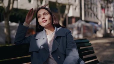 Şık mavi paltolu zarif genç bir kadın şehirdeki bir bankta düşünceli bir şekilde güneşli bir günün tadını çıkarıyor. Park Bench 'te oturan Hayatı Yansıtan Genç Kadın