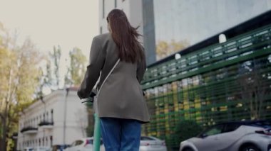 Kaygısız bir havası olan, ağaçlar ve modern arabalarla dolu bir sokak boyunca elektrikli scooter kullanan genç bir kadının arka planı. Bir kadın şehirde elektrikli scooterla keyifli bir yolculuğun tadını çıkarıyor.