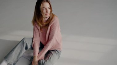 Rahat pembe bir kazak ve kot pantolon giyen düşünceli genç bir kadın zarif bir şekilde oturur, minimalist bir stüdyo ortamında yana bakar. Günlük Kıyafetli Genç Kadın Stüdyoda Düşünceli Poz Veriyor