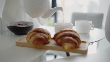 Ahşap bir tepside taze kruvasan ve modern bir mutfakta fincana çay dökülen sıcak bir kahvaltı sahnesi. Rahat sabah rutinlerini göstermek için mükemmel..