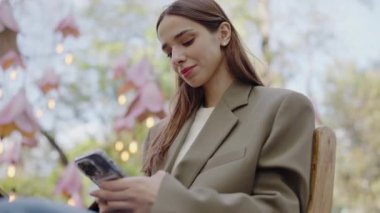 Zarif genç bir kadın, şık bir açık hava kafesinde otururken akıllı telefonuyla etkileşime girer. Etrafı hafif ışıklandırma ve yeşilliklerle çevrilidir. Açıkhava Kafe Ayarlarında Akıllı Telefon Kullanan Genç Kadın