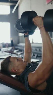 Salonda dambıllarla ağırlık kaldıran genç bir adam görülüyor. Odaklanmış ve kararlı. Spor salonunda halter kaldıran genç adam ağırlık kaldırma egzersizi yapıyor. Dikey