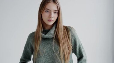 Yeşil kazaklı, katmanlı saçlı bir kadın, elleri kalçasında poz veriyor. Havalı bir etkinlikte mavi bir koluyla moda tasarımı sergiliyor.