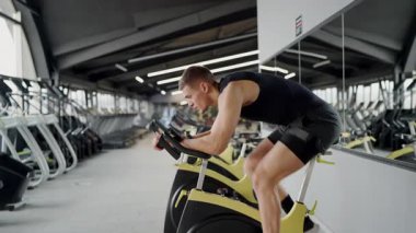 Tam teçhizatlı bir spor salonunda sabit bir spor bisikletine odaklanmış genç bir bisikletçi. Güç ve kararlılık sergiliyor. Spor salonunda kararlı genç adam sıkı çalışıyor.