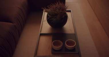 Sıcak, davet edici bir iç mekan. Ahşap sehpa, bitki süsü, küçük ahşap kaseler ve bir kitap. Rahat ve modern tasarım. Yumuşak, sıcak ışık. Rahatlamak için mükemmel bir ortam.