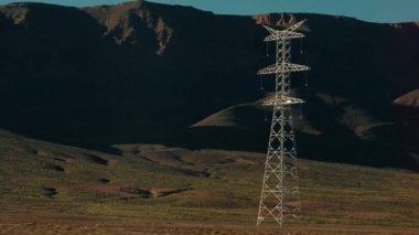 Atlas Dağları 'nın arka planında bir elektrik direği dik duruyor. Bereketli bitki örtüsü çöl zeminini kaplarken, kayalık tepeler dramatik bir şekilde arkalarında yükseliyor..