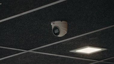 Koyu gri bir tavana beyaz bir güvenlik kamerası monte edilmiştir. Kamera aşağıya dönük. Tavanda kare ışık panelleri var. Bu düzenek güvenliği arttırır ve gözetleme sağlar..