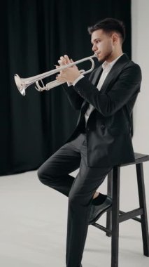 Takım elbiseli genç ve yakışıklı bir adam arzu ve ilhamla trompet çalar. Modern bir stüdyoda siyah ve beyaz arka planda trompet çalan bir erkek caz şarkıcısı. dikey video.