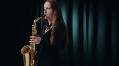 Siyah elbiseli genç bir kadın stüdyoda siyah arka planda saksafon çalıyor. Saksafon çalan caz müzisyeni. Siyah bir gece elbisesi giymiş, sahnede solo yapan bir kadın saksafoncu..