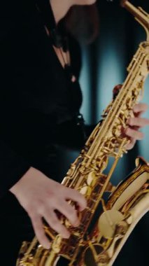 Siyah elbiseli genç bir kadın stüdyoda siyah arka planda saksafon çalıyor. Modern stüdyoda saksafon çalan bir kadın. Saksafon çalan caz müzisyeni. dikey