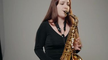 Siyah elbiseli genç bir kadın stüdyoda beyaz arka planda saksafon çalıyor. Siyah bir gece elbisesi giymiş, sahnede solo yapan bir kadın saksafoncu..
