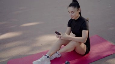Odaklanmış genç bir kadın parkta pembe bir yoga minderine oturur ve spor yaptıktan sonra telefonuna göz atar. Açık hava fitness ve teknoloji günlük rutinlere karışıyor. Park 'taki Yoga Mat' te akıllı telefon kullanan genç kadın.