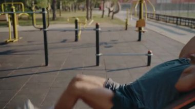 Formda bir genç adam, vücut kaslarını açık spor salonundaki eğimli bir bankta çalıştırıyor. Kararlılık ve güç göstererek fiziksel sağlığa ve dayanıklılığa odaklanır..