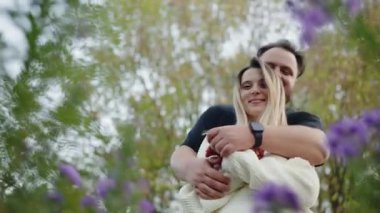 Doğayla çevrili romantik bir anın tadını çıkaran genç bir çiftin yürek ısıtan sahnesi. Adam sevgilisini sevgiyle öper, bulanık mor çiçekler ve ağaçlarla karakterize edilir..