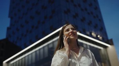 Genç bir kadın telefonuyla konuşurken gülümsüyor, arka planda modern binalarla neon ışığın altında duruyor. Şehirdeki Gece Telefonu Konuşmasından Hoşlanan Genç Kadın