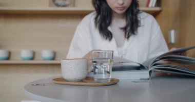 Odaklanmış genç bir kadın minimalist bir kafede parlak bir dergi okuyor. Masada seramik bir bardak, bir bardak su ve modern bir ahşap zemin var..